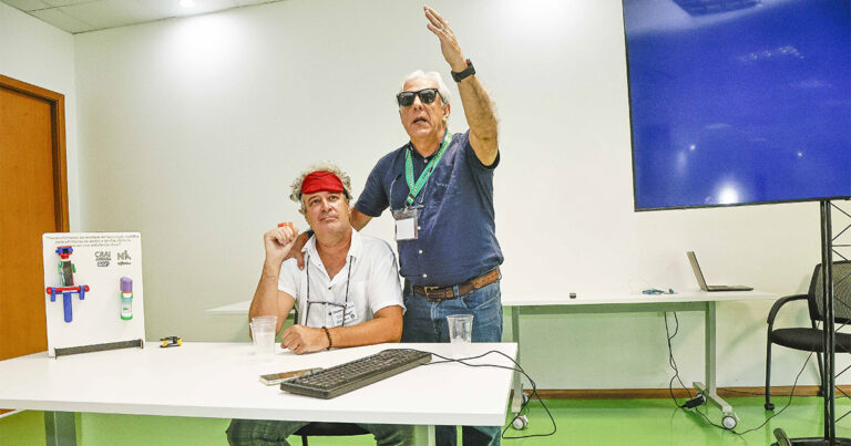 Um homem de óculos escuros em pé ao lado de um homem sentado com uma venda, demonstrando tecnologias assistivas.