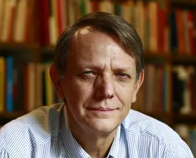 fotografia mostra André Singer em frente a uma estante de livros.