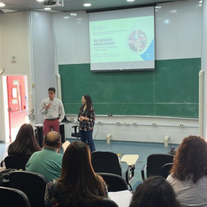 Imagem do professor Marcelo Nolasco e a Doutoranda Fernanda. Eles estão em pé em uma sala de aula. Ao fundo, há um quadro verde e uma projeção de Slide. Na foto é possível ver parte do público, pois o registro foi realizado na metade/fundo da sala.