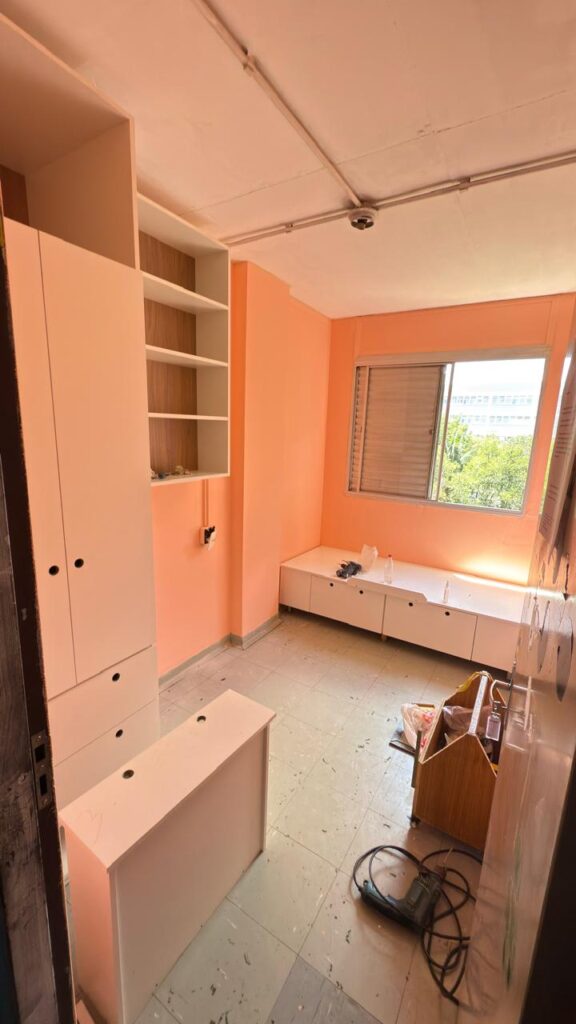 Imagem interna de quarto com paredes rosa, janela aberta com árvores ao fundo, cômodas brancas e armário.